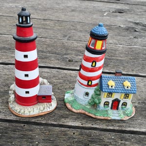 Può includere: Due statuine di faro in ceramica. Una è una torre dritta a strisce rosse e bianche con la parte superiore nera. L'altro è un faro inclinato con strisce rosse e bianche, un tetto blu e una casetta attaccata. Su una superficie di legno.