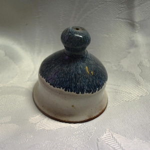 Puede incluir: Un tapón de botella de cerámica azul y blanco con la parte superior redondeada. El tapón tiene un esmalte azul moteado con un borde blanco.