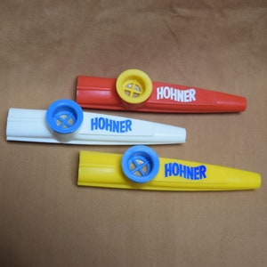 May include: Three plastic Hohner brand kazoos in red, white, and yellow. Each kazoo has a blue mouthpiece and a hole for blowing.