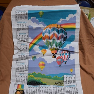 May include: A 1988 calendar towel with a rainbow and hot air balloons. The towel features a calendar for each month of the year. The towel is white with a blue background and a green border.