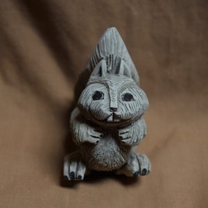 May include: A gray wooden squirrel figurine with a bushy tail, black eyes, and a small smile. The figurine is standing on a brown surface.