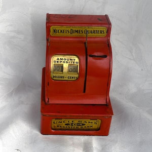 May include: A vintage red metal Uncle Sam's 3 Coin Register Bank with gold-toned labels. The top label reads Nickels Dimes Quarters, and the front display shows Amount Deposited in Dollars and Cents. The base features the text Uncle Sam's 3 Coin Register Bank.