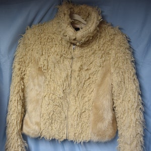 May include: A cream-coloured faux fur jacket with a zip fastening and a high collar. The jacket has a shaggy texture and is lined with a smooth, cream-coloured fabric.