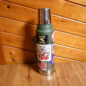 May include: A green and silver insulated thermos with a stainless steel lid. The thermos has several stickers, including one with a bulldog and the words "Big Dog 106" and another with an American flag.