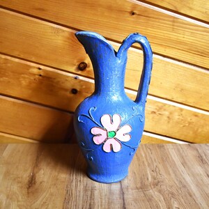 May include: A blue ceramic pitcher with a handle and a decorative flower design. The flower is pink with a green center and is surrounded by swirling vines. The pitcher is on a wooden surface.