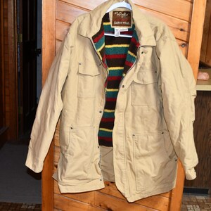 Op de afbeelding: Een beige jas met een kleurrijke gestreepte voering, hangend aan een houten deur. De jas heeft meerdere zakken en de merknaam "Walls Ranch Wear" is zichtbaar op het label.