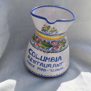 May include: A white ceramic pitcher with a blue rim, featuring hand-painted floral motifs in green and pink, a yellow band, and blue text that reads SANGRIA, COLUMBIA RESTAURANT, SINCE 1905-FLORIDA, with a decorative scalloped blue line at the base.