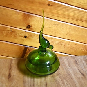 May include: A translucent emerald green glass jar with a rounded base and a matching lid. The lid features a tall, slender, curved finial that tapers to a point, resembling a stylized bird or abstract organic form, creating a smooth, polished finish.