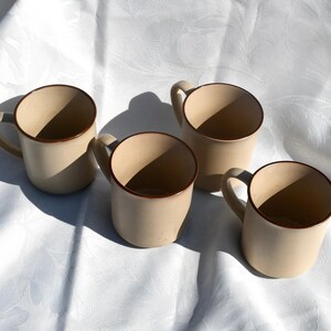 May include: Four beige ceramic mugs with brown trim and handles are arranged on a white, textured surface. The mugs are evenly spaced, with shadows cast across the surface. The mugs are empty.