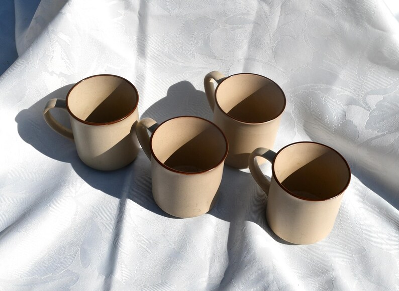 May include: Four beige ceramic mugs with brown trim and handles are arranged on a white, textured surface. The mugs are evenly spaced, with shadows cast across the surface. The mugs are empty.