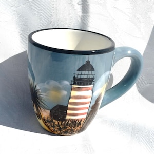 May include: A ceramic mug with a light blue exterior and a white interior. The mug features a painted scene of a lighthouse with red and white horizontal stripes, a black lantern room, and dark silhouettes of coastal vegetation and clouds.