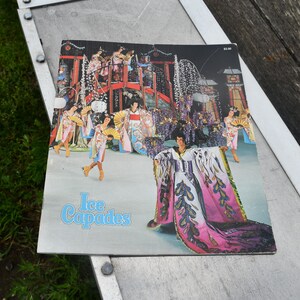 May include: A vintage Ice Capades program cover featuring a colorful illustration of a Japanese-themed ice show. The image showcases performers in elaborate costumes, with a prominent figure in a flowing, pink and purple gown. The text "Ice Capades" is in blue.