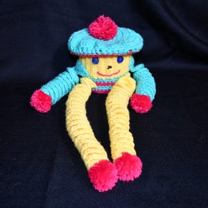 May include: A crocheted toy with a blue and yellow striped hat, yellow legs, and pink pom-poms. The toy has a smiling face with blue eyes.