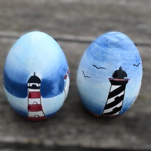 May include: Two painted egg-shaped ornaments. One is light blue with a red and white lighthouse. The other is dark blue with a black and white lighthouse and flying birds. The background is a wooden surface.