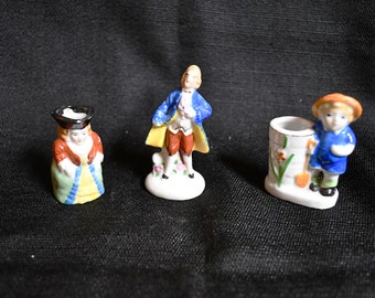 Puede incluir: Tres pequeñas figuritas de porcelana. Una mujer con un vestido verde, amarillo y rojo con un sombrero negro, un hombre con un abrigo azul y pantalones rojos, y un niño con un abrigo azul y un sombrero naranja sosteniendo una pala.