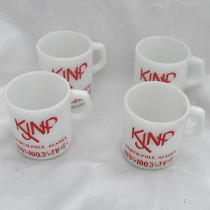 Può includere: Quattro tazze in ceramica bianca con scritte rosse e la scritta "KNP NORTH POLE, ALASKA 170 100.3 TV". Ogni tazza ha un piccolo manico e una forma cilindrica. Le tazze sono disposte su una superficie bianca.