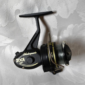 May include: Black Shakespeare 2001i fishing reel with gold accents. The reel has a handle, a spool with fishing line, and the text "Shakespeare" and "2001i" in gold lettering. The reel is designed for fishing.