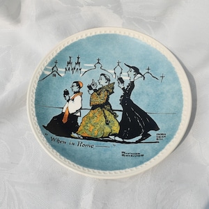 May include: A decorative plate with a light blue background and a white rim. The plate features an illustration of three figures in religious attire, with the text "When in Rome." The artist's signature is visible.