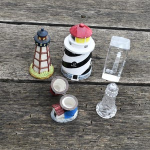 Puede incluir: Una colección de figuras de faros en miniatura de varios diseños. Uno es una estructura transparente similar al vidrio, otro tiene rayas blancas y negras con una parte superior roja, y otros están pintados con detalles. Los objetos están dispuestos sobre una superficie de madera envejecida.