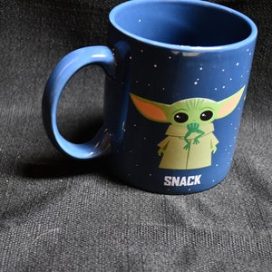 May include: A blue ceramic mug featuring a cartoon character. The character is green with large eyes and is holding a frog in its mouth. The mug has the word "SNACK" printed on the bottom.