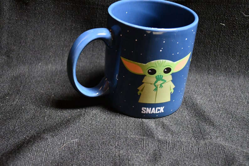May include: A blue ceramic mug featuring a cartoon character. The character is green with large eyes and is holding a frog in its mouth. The mug has the word "SNACK" printed on the bottom.