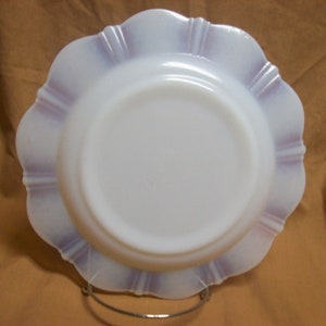 May include: A white milk glass plate with a scalloped edge and a lavender iridescent design. The plate is on a brown surface.