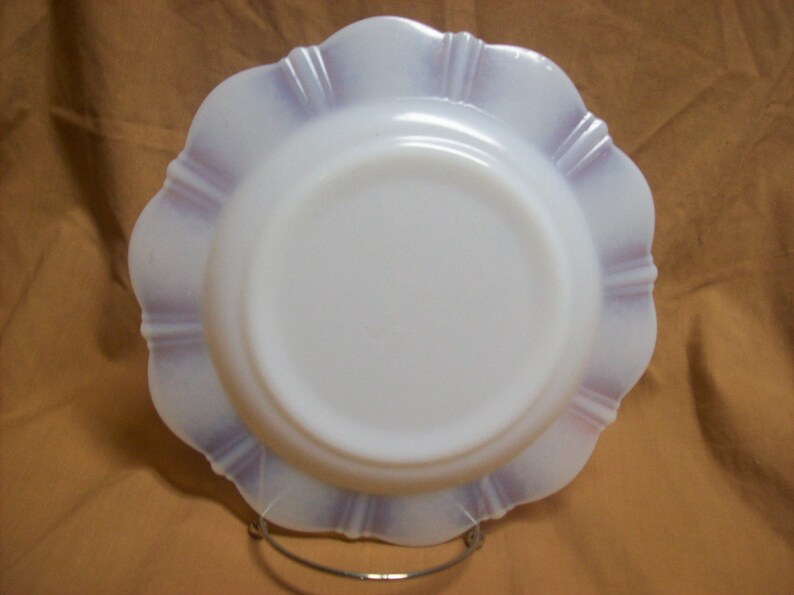 May include: A white milk glass plate with a scalloped edge and a lavender iridescent design. The plate is on a brown surface.