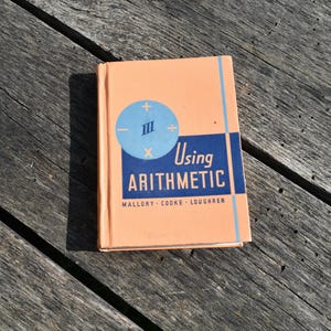 Puede incluir: Un libro de texto antiguo titulado "Using Arithmetic" con una cubierta de color melocotón y un diseño geométrico azul y blanco. La portada del libro presenta símbolos matemáticos y los nombres de los autores.