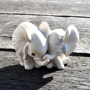 May include: A white ceramic figurine of two elephants embracing. The elephants have detailed features, including large ears and trunks intertwined. One elephant has a small floral accent. The figurine is on a weathered wooden surface.