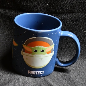 May include: A blue ceramic mug with a white and orange spaceship design. The spaceship has a small green creature inside. The text "PROTECT" is printed on the bottom of the mug.