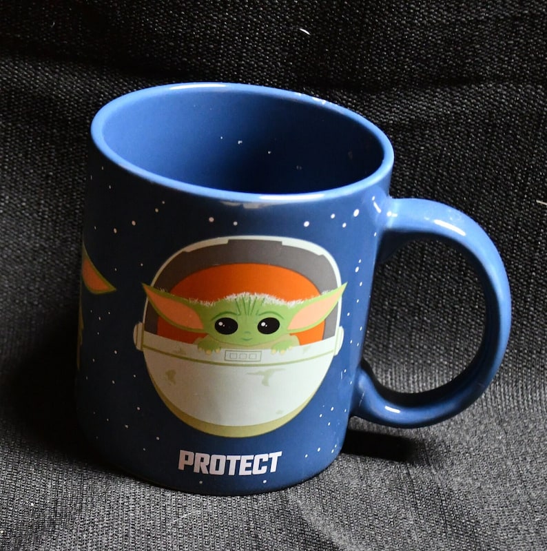 May include: A blue ceramic mug with a white and orange spaceship design. The spaceship has a small green creature inside. The text "PROTECT" is printed on the bottom of the mug.