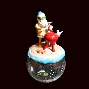 Op de afbeelding: Een decoratieve glazen pot met een strandthema. De pot heeft een miniatuurfiguur van een man die grilt op een rode barbecue, een krab en een zeemeeuw. De pot heeft een helder, getextureerd oppervlak met een groene visillustratie.