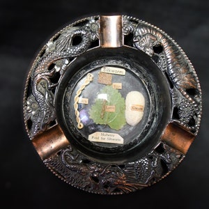May include: A round, copper and silver ashtray with a dragon design. The center of the ashtray has a glass dome with a silk worm, cocoon, and mulberry leaf inside. The text "Silkworm", "Egg", "Larva", "Cocoon", "Mulberry Food for Silkworm" is printed on the labels inside the dome.