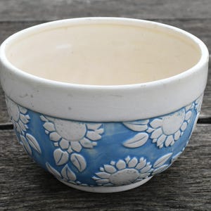 May include: A ceramic bowl featuring a smooth white upper rim and a textured lower section decorated with embossed white sunflower patterns against a blue background. The interior of the bowl is a solid cream color with a visible crackle glaze finish.