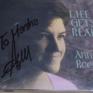 Puede incluir: Un CD firmado por Ann Reed, titulado "Life Gets Real". La portada del CD presenta un primer plano de una mujer con cabello castaño corto, vistiendo una camisa rosa. La firma está en tinta negra y dice "To Marsha".