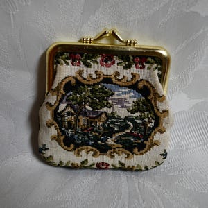 Puede incluir: Una pequeña cartera de monedas de tela blanca con un marco de metal dorado. La cartera tiene un diseño floral y una escena bordada de una casa y árboles en la parte delantera.
