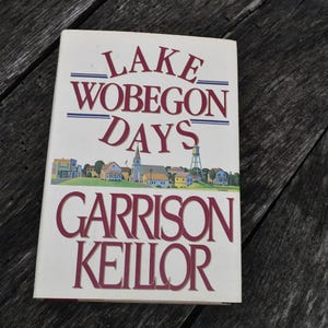 May include: A hardcover book titled Lake Wobegon Days by Garrison Keillor. The white cover features a colorful illustration of a small town with houses, a church, and a water tower. The title and author name are printed in large red serif font.