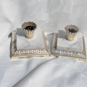 May include: Two matching antique candle holders. Each has a square marble base with a gold-colored metal frame. The candle cups are gold-colored and have a scalloped edge. The bases are white and gray.