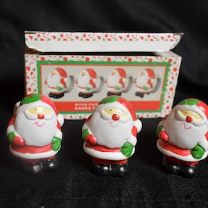 May include: Set of four ceramic Santa Claus napkin rings. Each ring features a red and white Santa Claus with a green hat and a white beard. The rings are packaged in a box with a festive design.