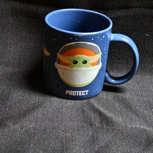 May include: A blue ceramic mug featuring a white and orange graphic of a character from the Star Wars franchise. The character is in a spaceship with the text "PROTECT" below it.
