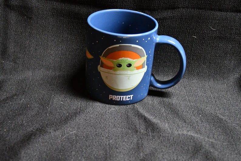 May include: A blue ceramic mug featuring a white and orange graphic of a character from the Star Wars franchise. The character is in a spaceship with the text "PROTECT" below it.