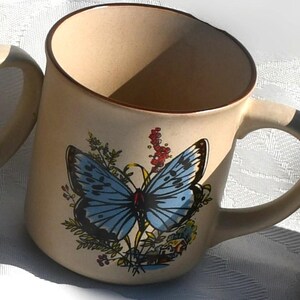 May include: Beige ceramic mug featuring a blue butterfly design with black and white accents. The mug has a brown rim and handle, and is decorated with floral elements. The mug is a standard size for coffee or tea.