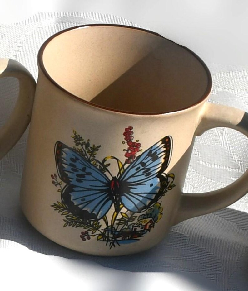 May include: Beige ceramic mug featuring a blue butterfly design with black and white accents. The mug has a brown rim and handle, and is decorated with floral elements. The mug is a standard size for coffee or tea.