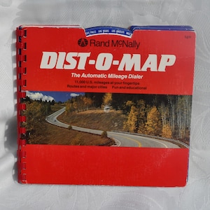 May include: A red Rand McNally DIST-O-MAP, an automatic mileage dialer, with a spiral binding. The cover features a scenic road winding through a forest. Text includes "11,000 U.S. mileages at your fingertips" and "Routes and major cities."