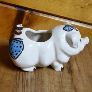 May include: A white ceramic pig-shaped planter with a large opening. The pig has blue strawberry designs with black dots on its side and top. The planter is on a wooden surface.