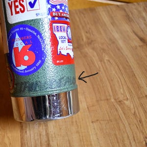 May include: A cylindrical metal object with a green textured body and a shiny silver base. The object has several stickers, including one for Local Union 16, Omaha, Nebraska, and another with the text "BUY AMERICAN".