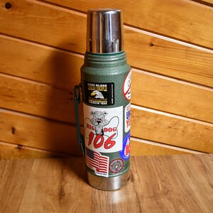 May include: A green insulated thermos with a stainless steel lid and base. The thermos is adorned with various stickers, including one with a bulldog graphic and the text "BIG DOG 106". Other stickers feature patriotic themes and text.