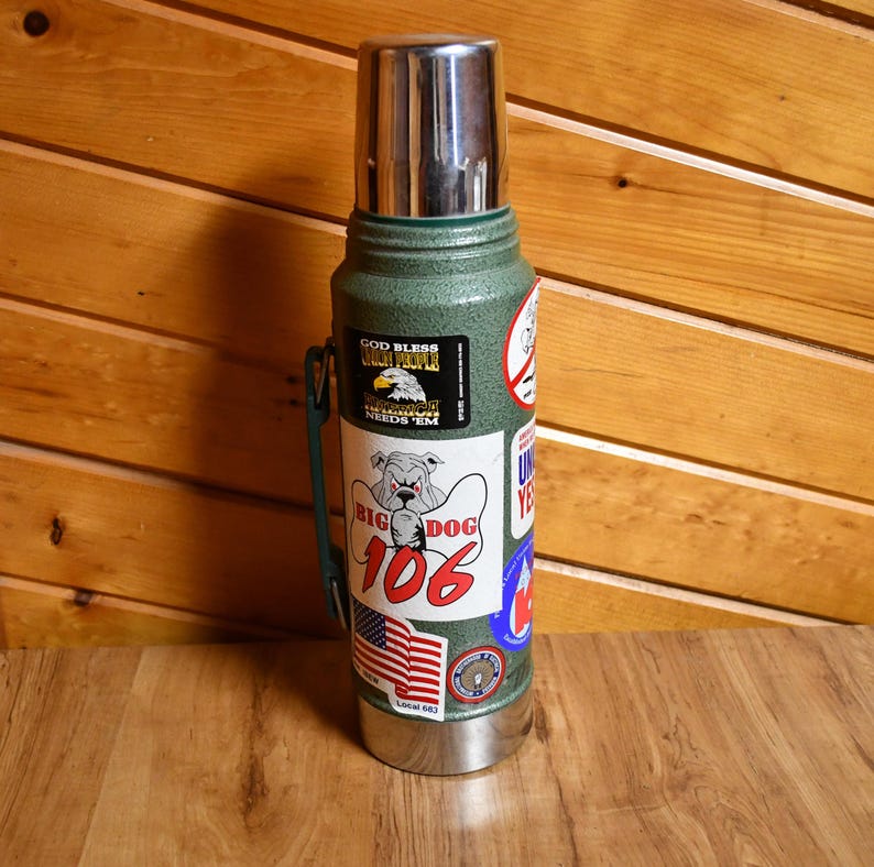 May include: A green insulated thermos with a stainless steel lid and base. The thermos is adorned with various stickers, including one with a bulldog graphic and the text "BIG DOG 106". Other stickers feature patriotic themes and text.