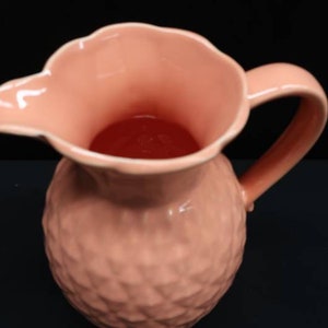 May include: A pink ceramic pitcher with a textured surface and a wide mouth. The pitcher has a handle and is designed for pouring liquids.