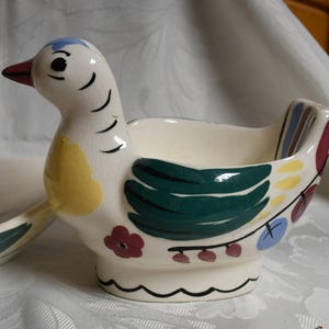May include: A white ceramic bird-shaped dish with green, yellow, red, and blue painted details. The dish has a small bowl on the back for holding eggs or other small items.